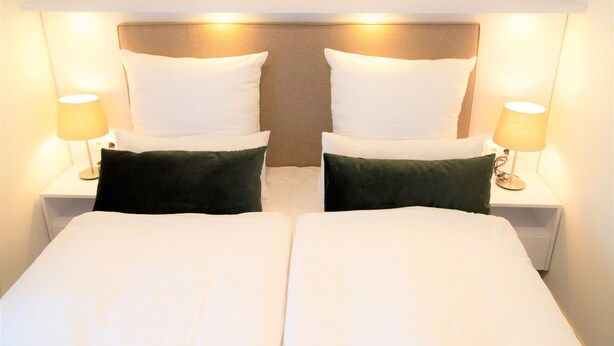 A double bed with white linens and two green pillows, flanked by two bedside tables with lamps, in a hotel room. - Amplidi Hotel Hannover (Hannover)