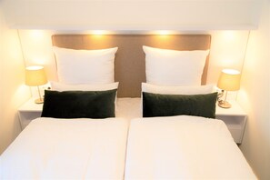 Una cama doble con sábanas blancas y dos almohadas verdes, flanqueada por dos mesitas de noche con lámparas, en una habitación de hotel.