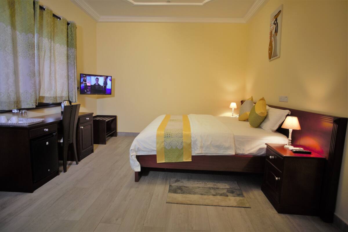 Quarto de hotel com cama, mesa de cabeceira, luminária, TV, escrivaninha, cadeira e um pequeno armário.