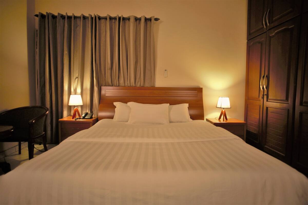 Quarto de hotel com uma cama grande, duas luminárias de cabeceira, uma cadeira e um guarda-roupa.