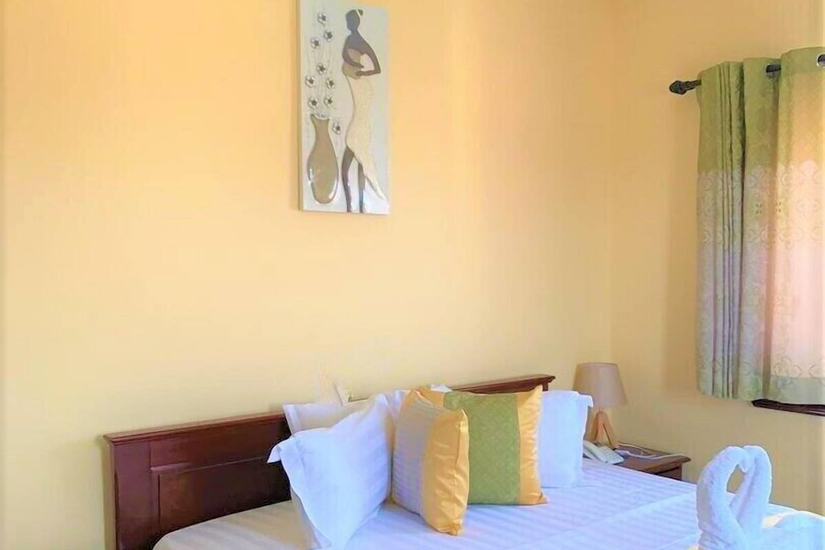 Quarto de hotel com cama, almofadas, mesa de cabeceira, luminária, um quadro na parede e janela com cortinas.
