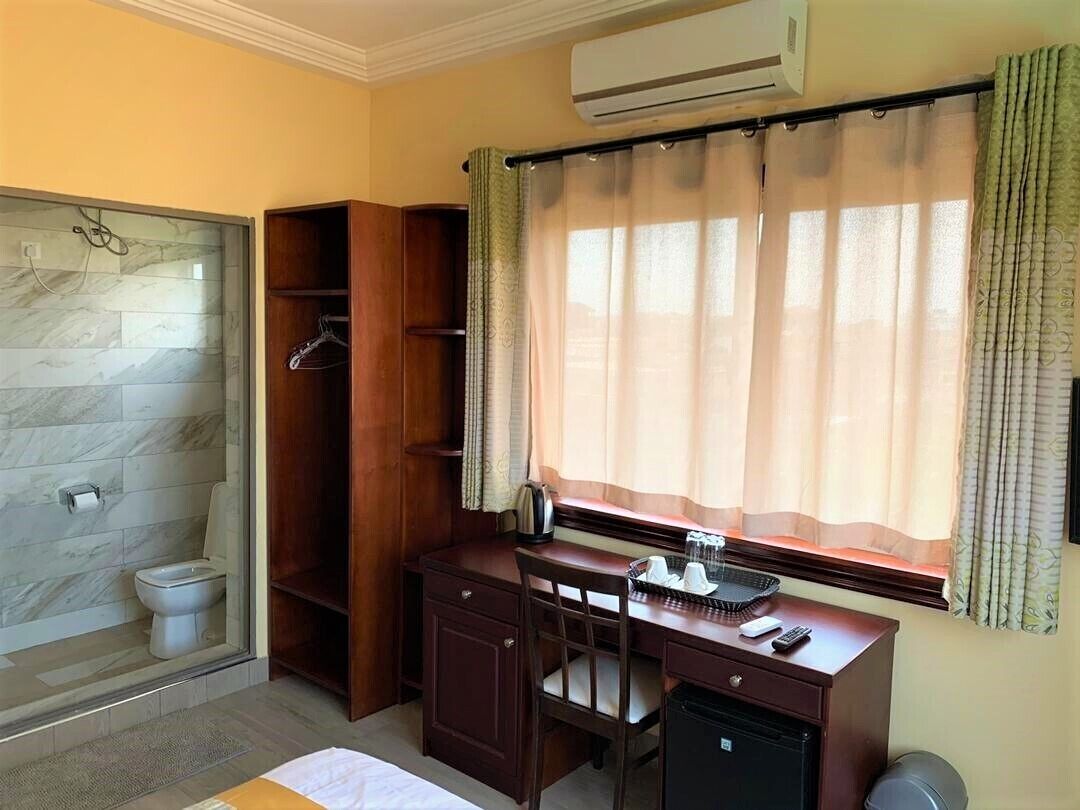 A compact hotel room with a shower, toilet, wooden desk, and a window with curtains.