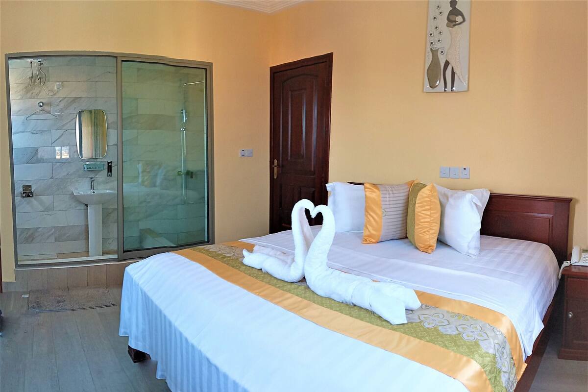 Quarto de hotel com cama, porta de madeira, box de vidro e uma obra de arte na parede.