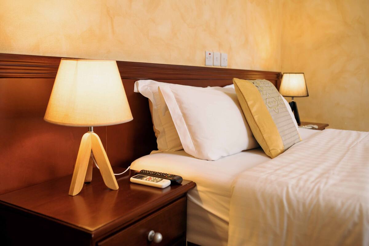 Uma cama bem arrumada com roupa de cama branca e bege, uma mesinha de cabeceira de madeira com uma luminária e um controle remoto ao lado.