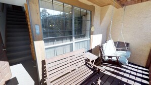 Un banco de madera y una silla mecedora en un espacio exterior cubierto, con piso a cuadros, una ventana con persianas y una escalera que conduce a un nivel superior.