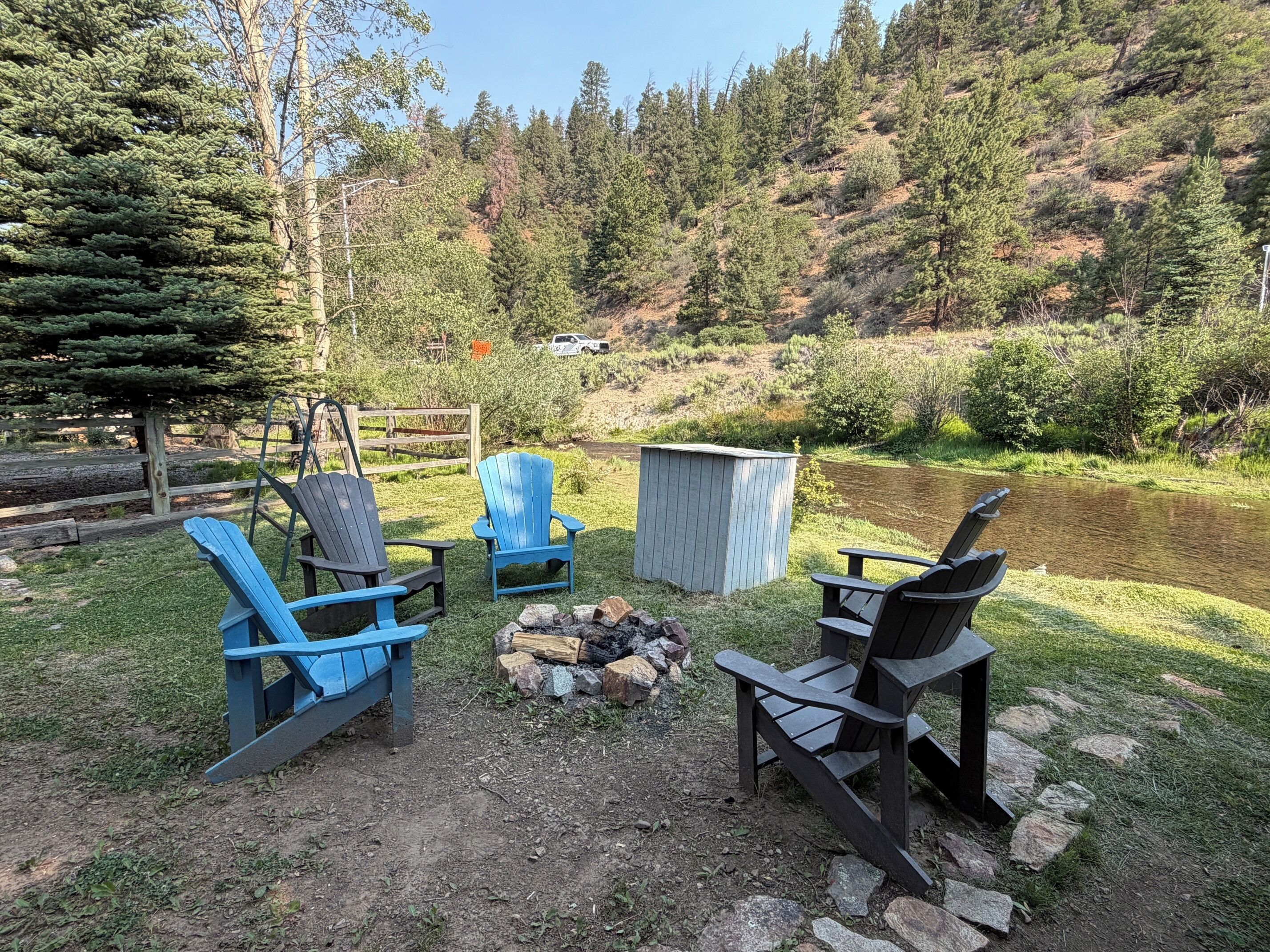 A campsite with four Adirondack chairs, a fire pit, and a shed, surrounded by a pond and forested hills.