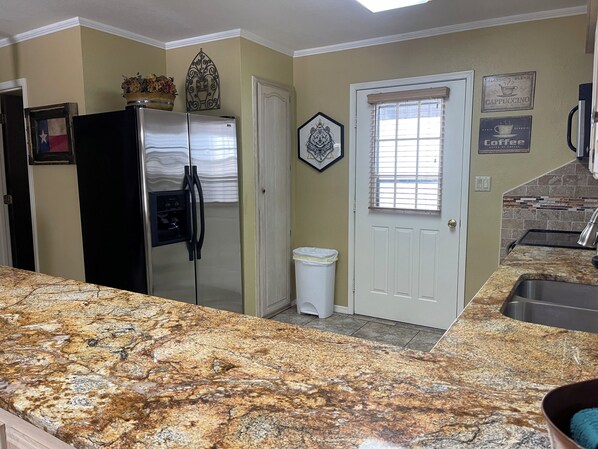 A kitchen with granite countertops, a stainless steel refrigerator, a white door, and a window with blinds.