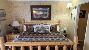Un lit en bois, recouvert d’une couvre-lit à motifs et agrémenté d’un coussin sur lequel est brodé le mot « AVENTURE ».