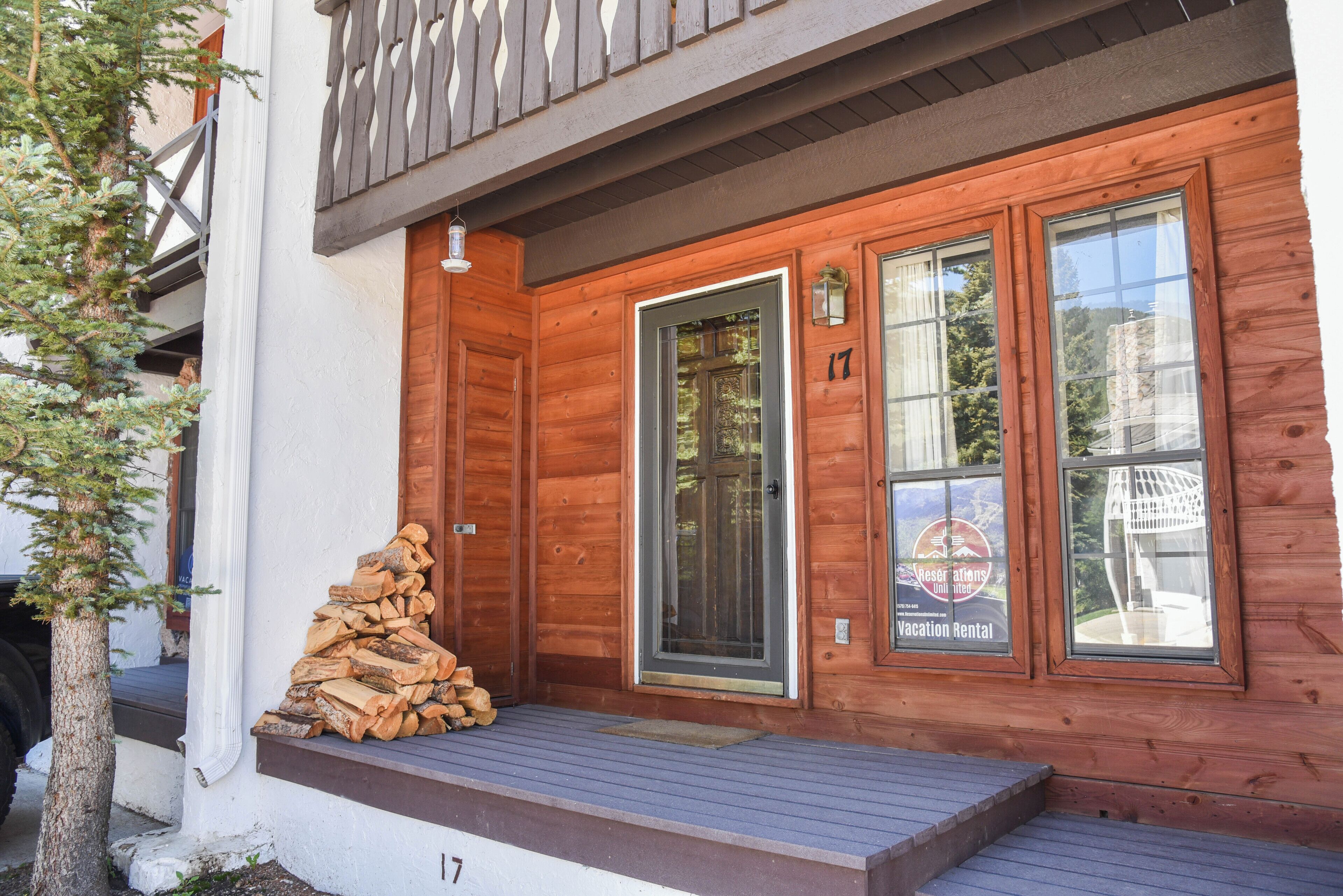 A wooden cabin with a front door, two windows, and a porch with a stack of firewood.