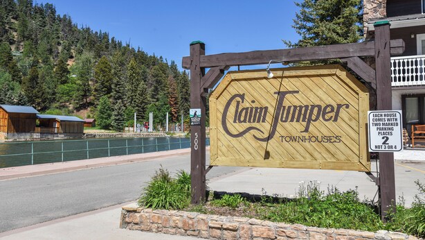 A wooden sign for Claim Jumper Townhouses with a clear view of the surrounding area including a road, a body of water, and a forested hillside. - Claim Jumper #17 Townhouse (Red River)