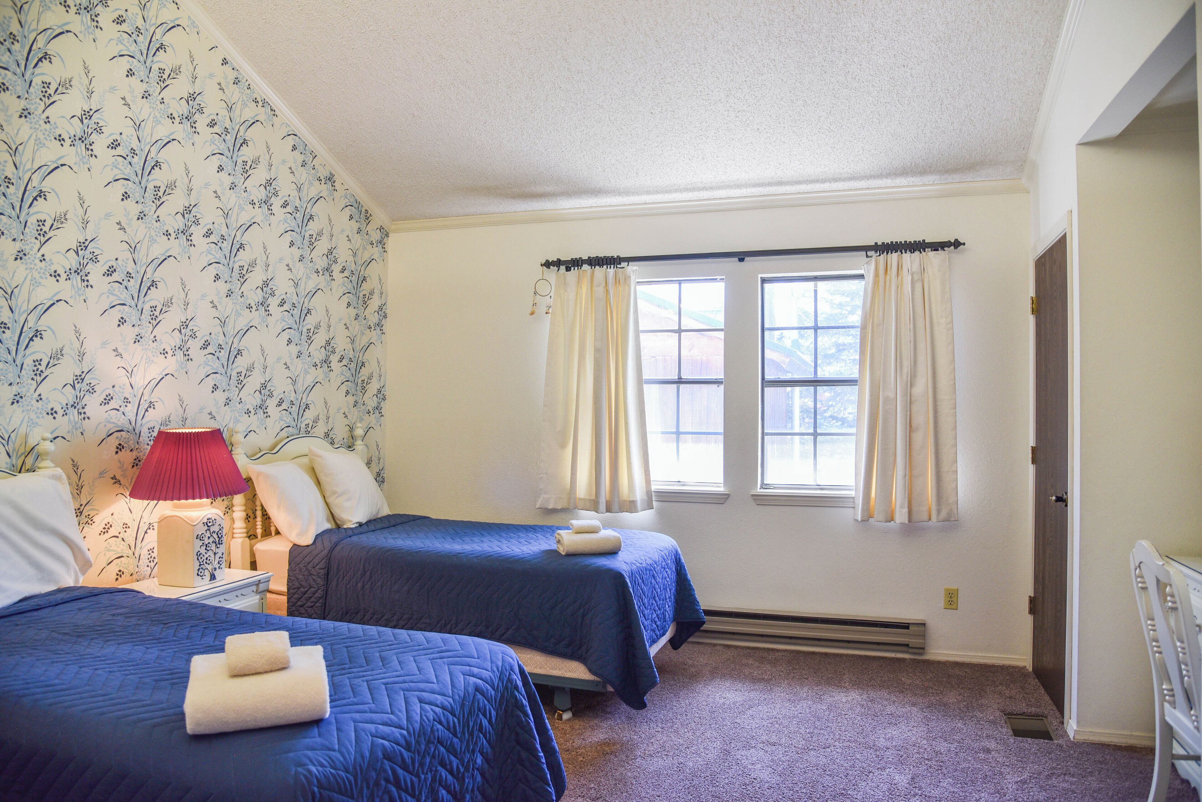 A hotel room with two beds, a nightstand, a lamp, and a window with curtains.