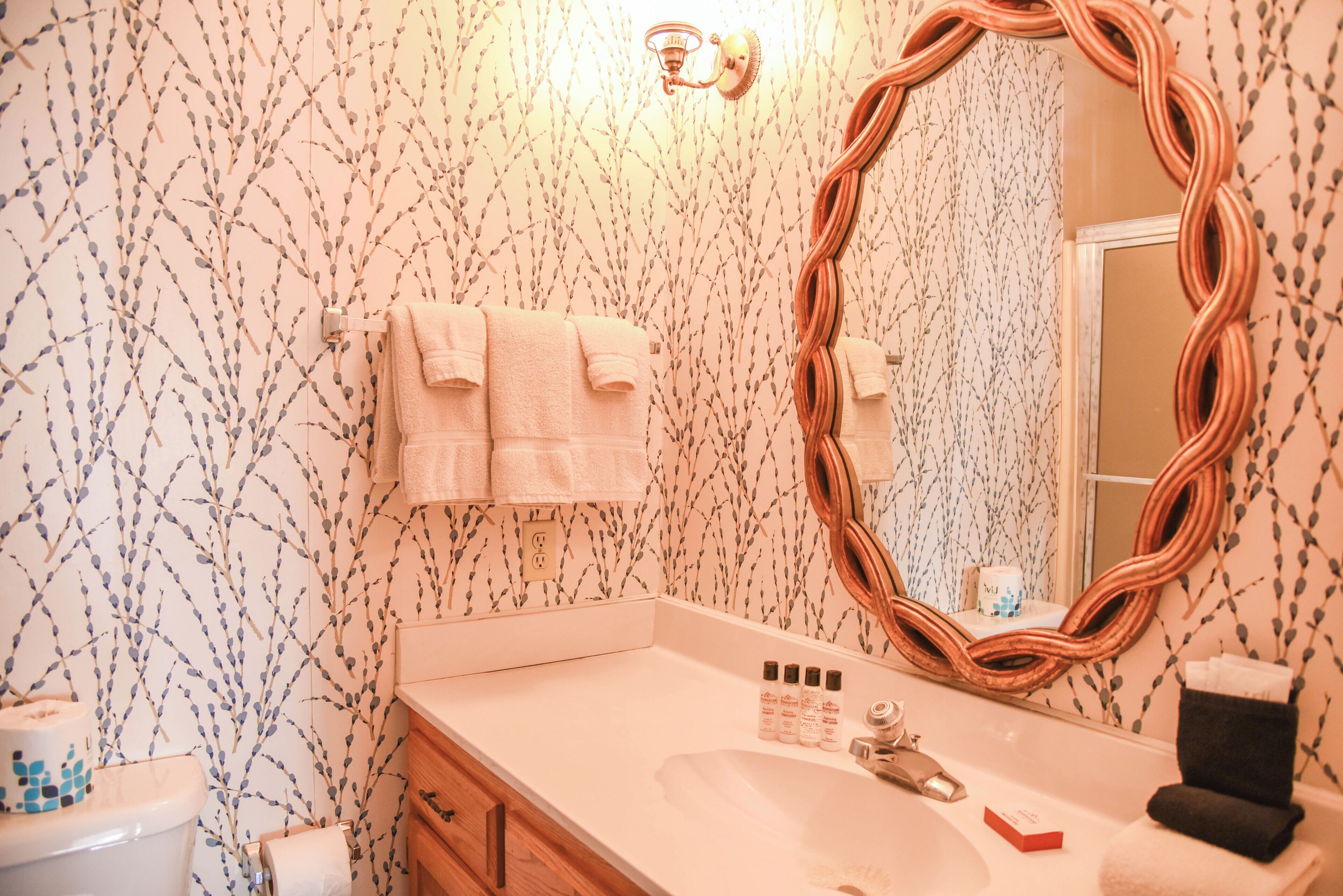 A bathroom with a white sink, a mirror, and a toilet. There are towels hanging on the wall and a bottle of shampoo on the sink.