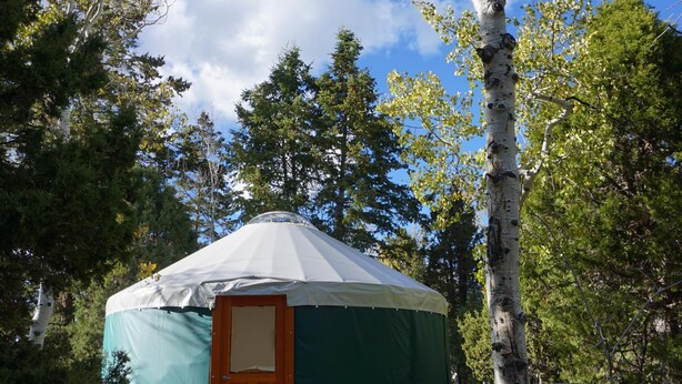 A yurt with a wooden door, surrounded by trees and under a clear sky. - Snowshoe or ski and relax by a warm wood stove while playing games! (Scofield)