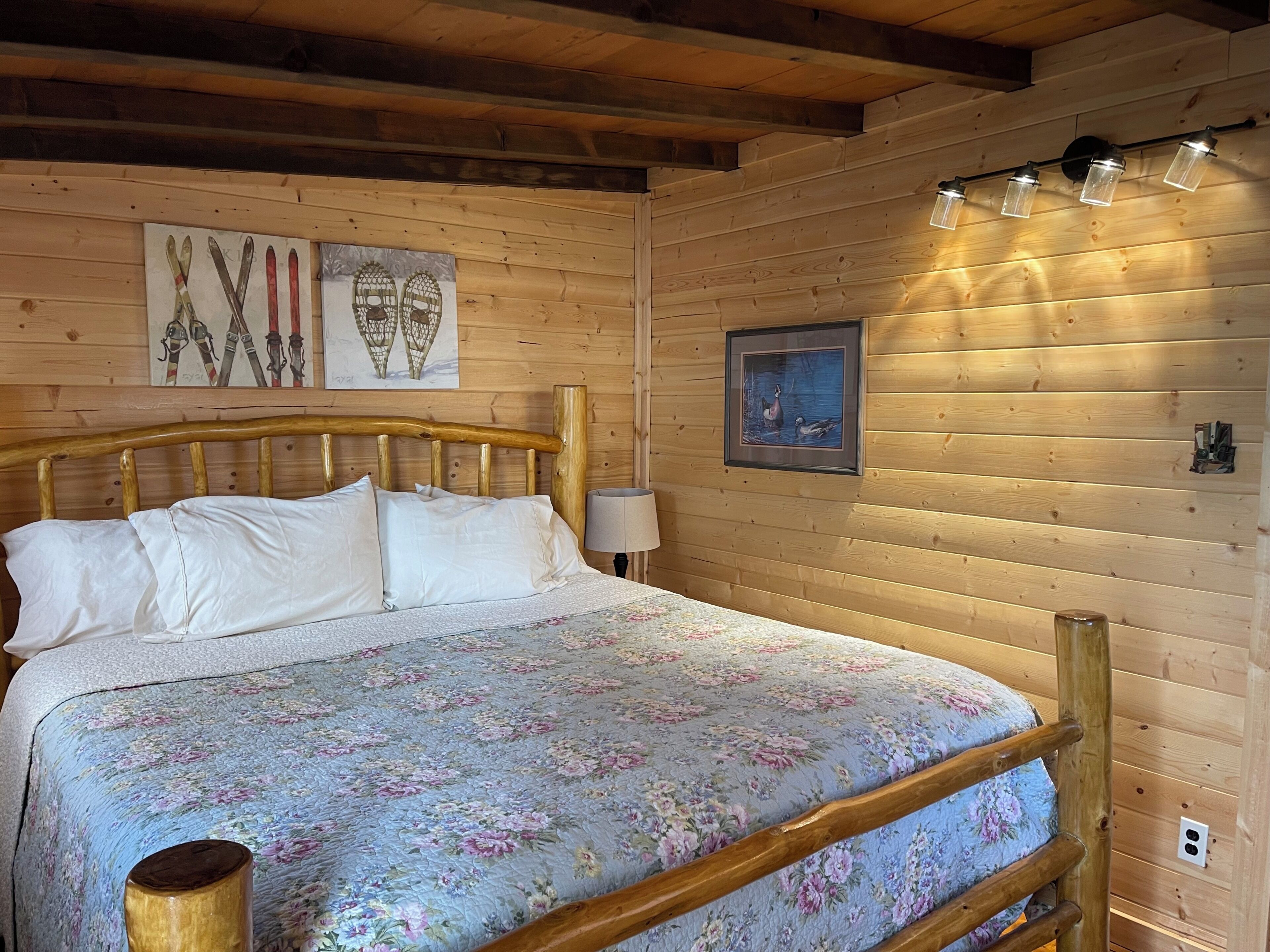 A wooden cabin bedroom with a bed, wooden headboard, wall-mounted light fixtures, and framed artwork.