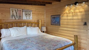 Une chambre dans une cabane en bois, avec un lit, une tête de lit en bois, des luminaires fixés au mur et des œuvres d’art encadrées.
