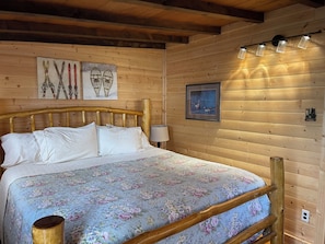 A wooden cabin bedroom with a bed, wooden headboard, wall-mounted light fixtures, and framed artwork.