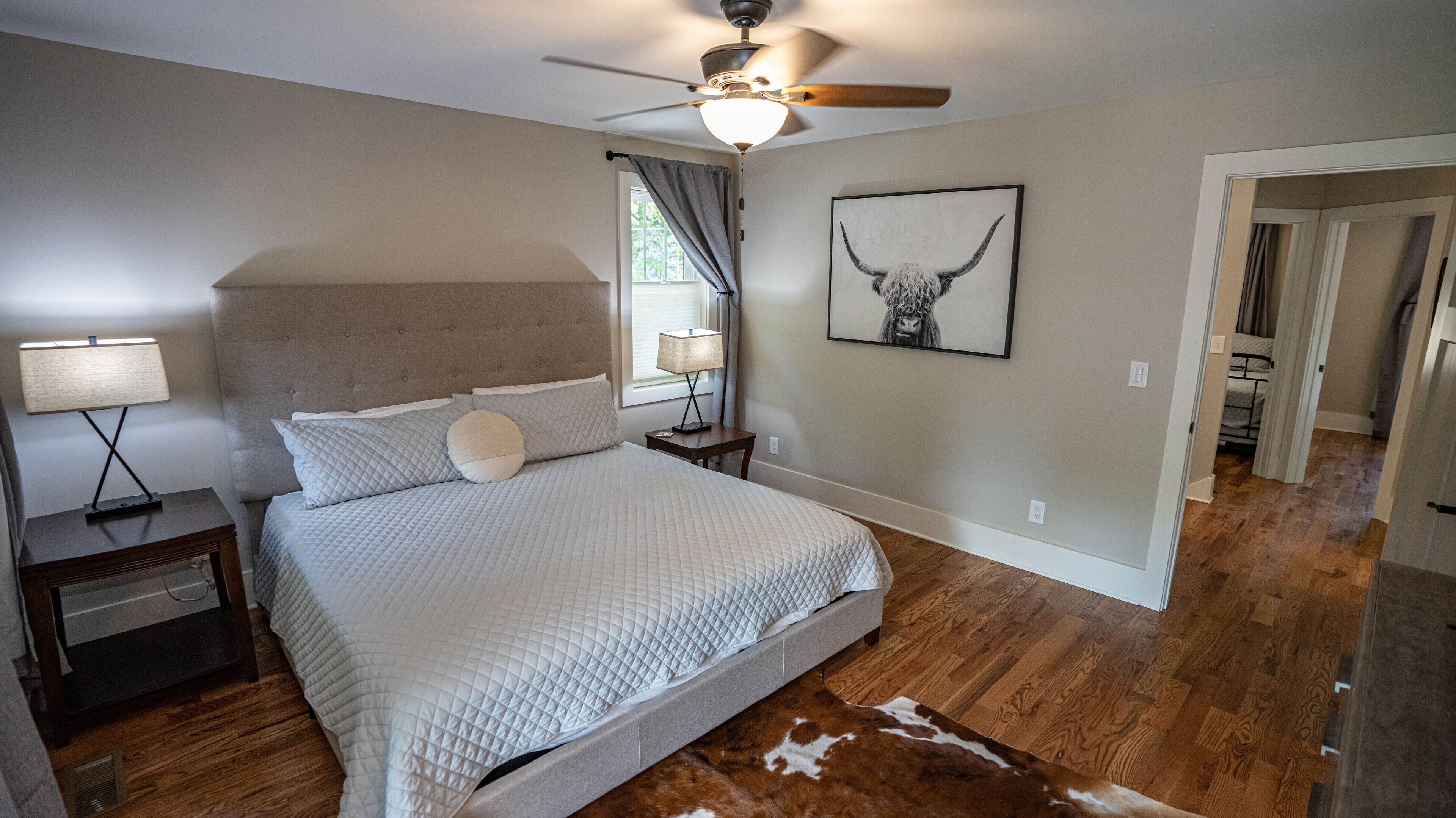 A bedroom with a bed, a nightstand, a lamp, a ceiling fan, a window with curtains, a wall-mounted picture, and a wooden floor.
