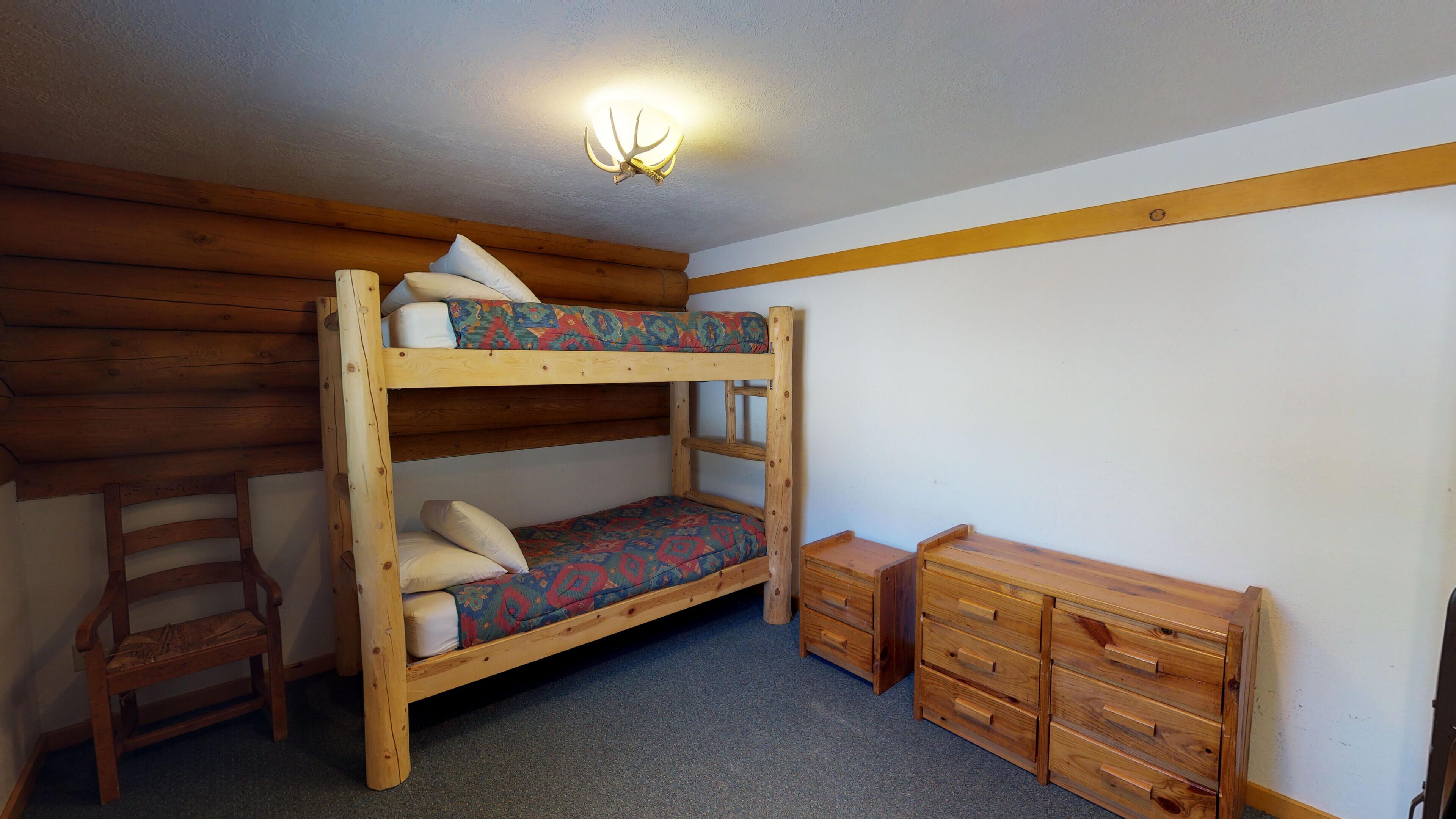A bunk bed with a patterned mattress, a wooden chair, a small wooden dresser, and a ceiling light fixture.