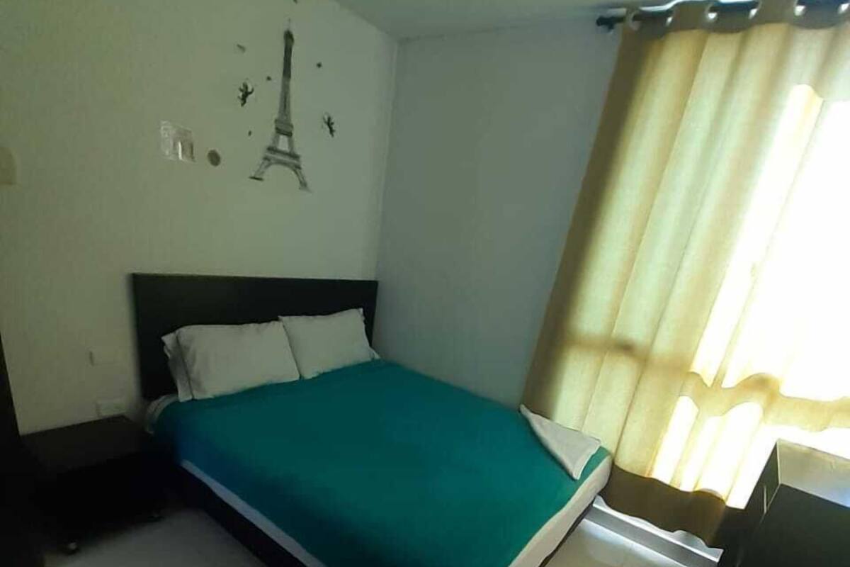 Un dormitorio con cama, mesita de noche, escritorio, silla, cortina y un adorno en la pared.