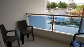 En balkon med to sorte plastikstole, gelænder af glas og udsigt til en blå bygning og gade.