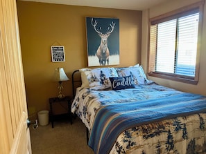 Une chambre à coucher avec un lit, une table de chevet, une lampe, un tableau encadré représentant un cerf, une pancarte et une fenêtre.