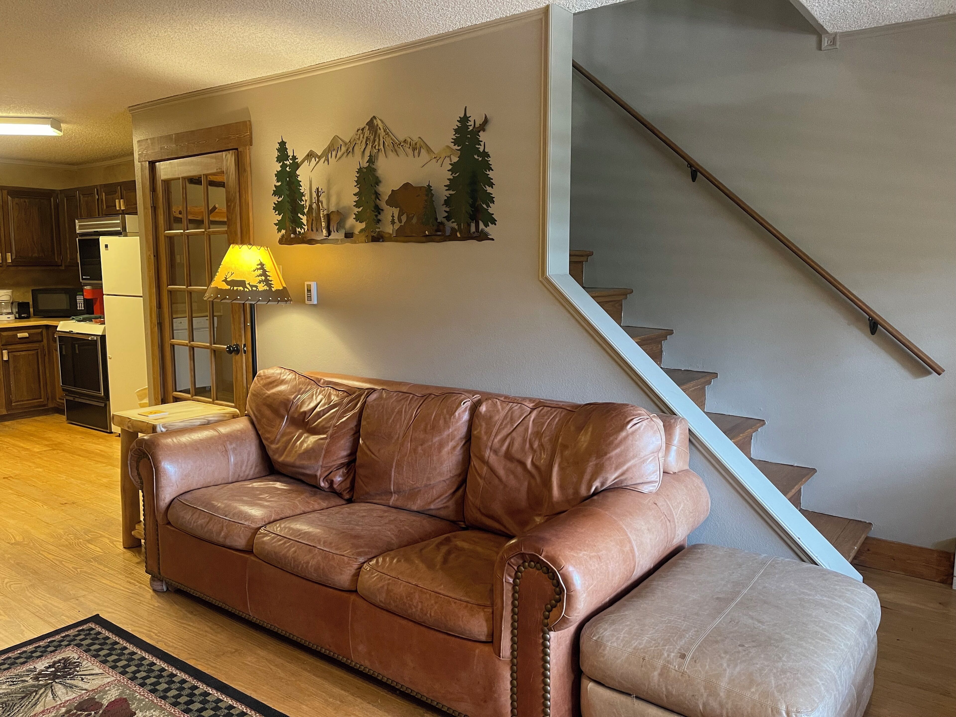 Un canapé en cuir marron dans un salon doté d’un plancher en bois, une cuisine à l’arrière-plan et un escalier sur la droite.