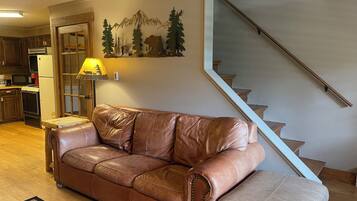 Un canapé en cuir marron dans un salon doté d’un plancher en bois, une cuisine à l’arrière-plan et un escalier sur la droite.