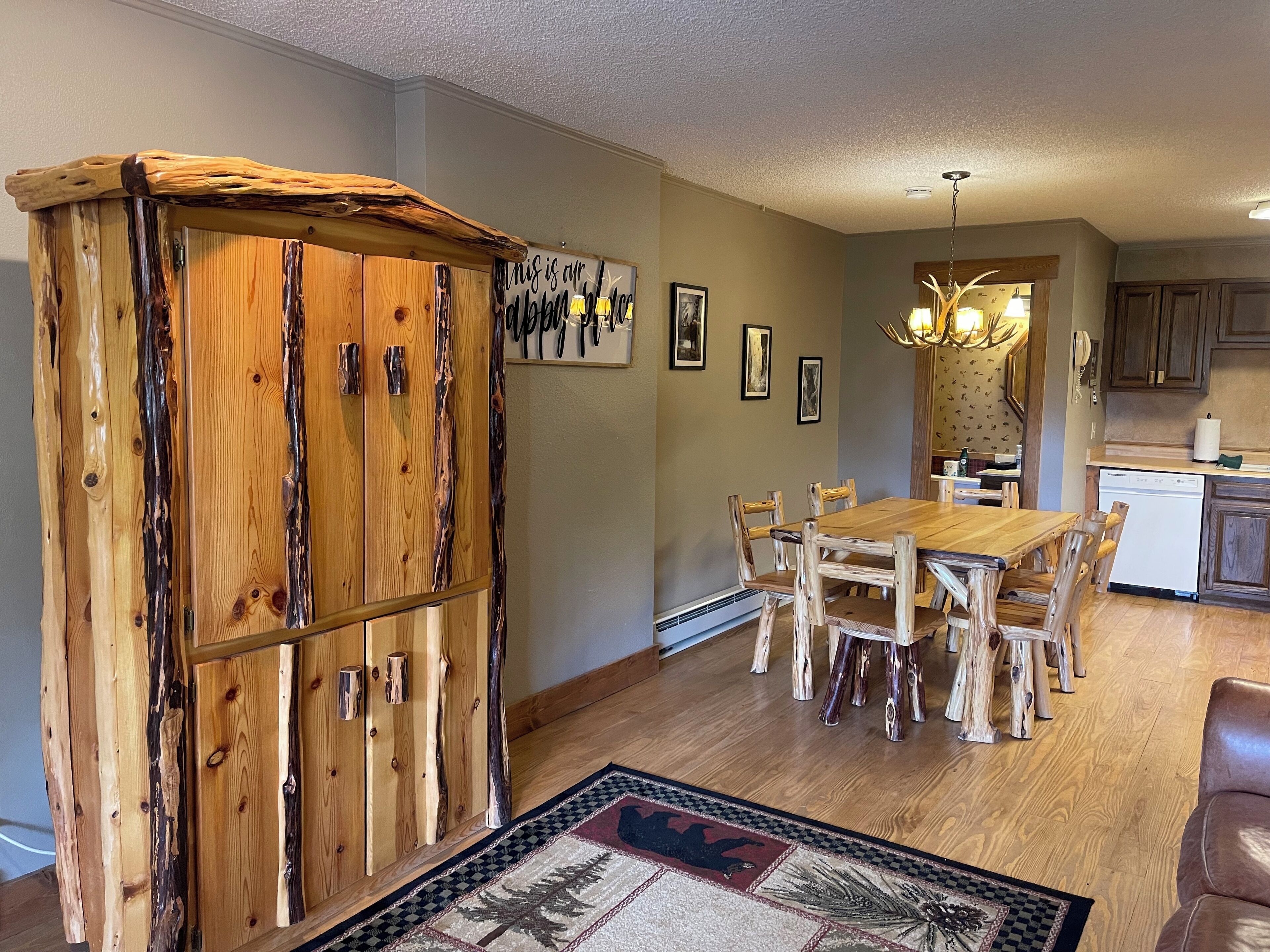 A dining area with a wooden table and chairs, a kitchen in the background, and a decorative rug on the floor.