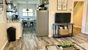 Una cocina moderna con comedor, una sala de estar con televisor y mesa de centro, y una escalera que sube a otro nivel.