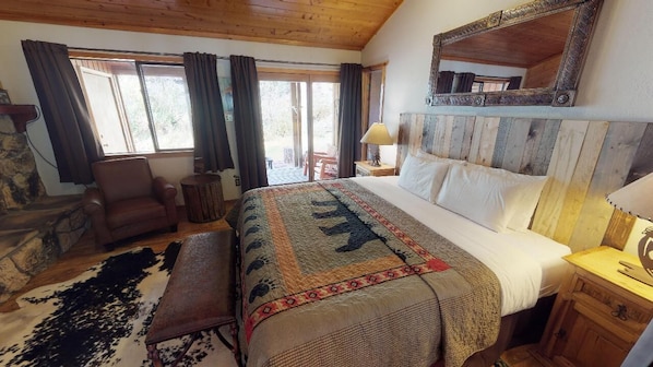 Un dormitorio con una cama grande, cabecera de madera, pared de piedra, una silla, una lámpara y una ventana con cortinas.