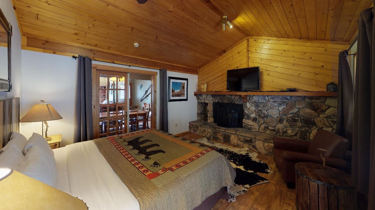 Interior de una cabaña de troncos con chimenea de piedra, una cama con manta estampada, un televisor montado sobre la chimenea y un sillón de cuero.
