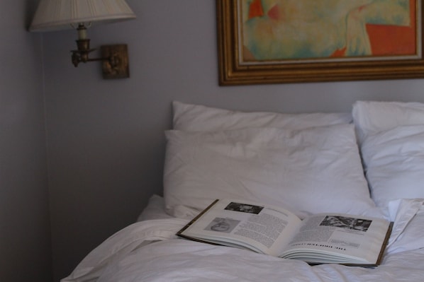 A neatly made bed with a book open on it, a lamp, and a painting on the wall.