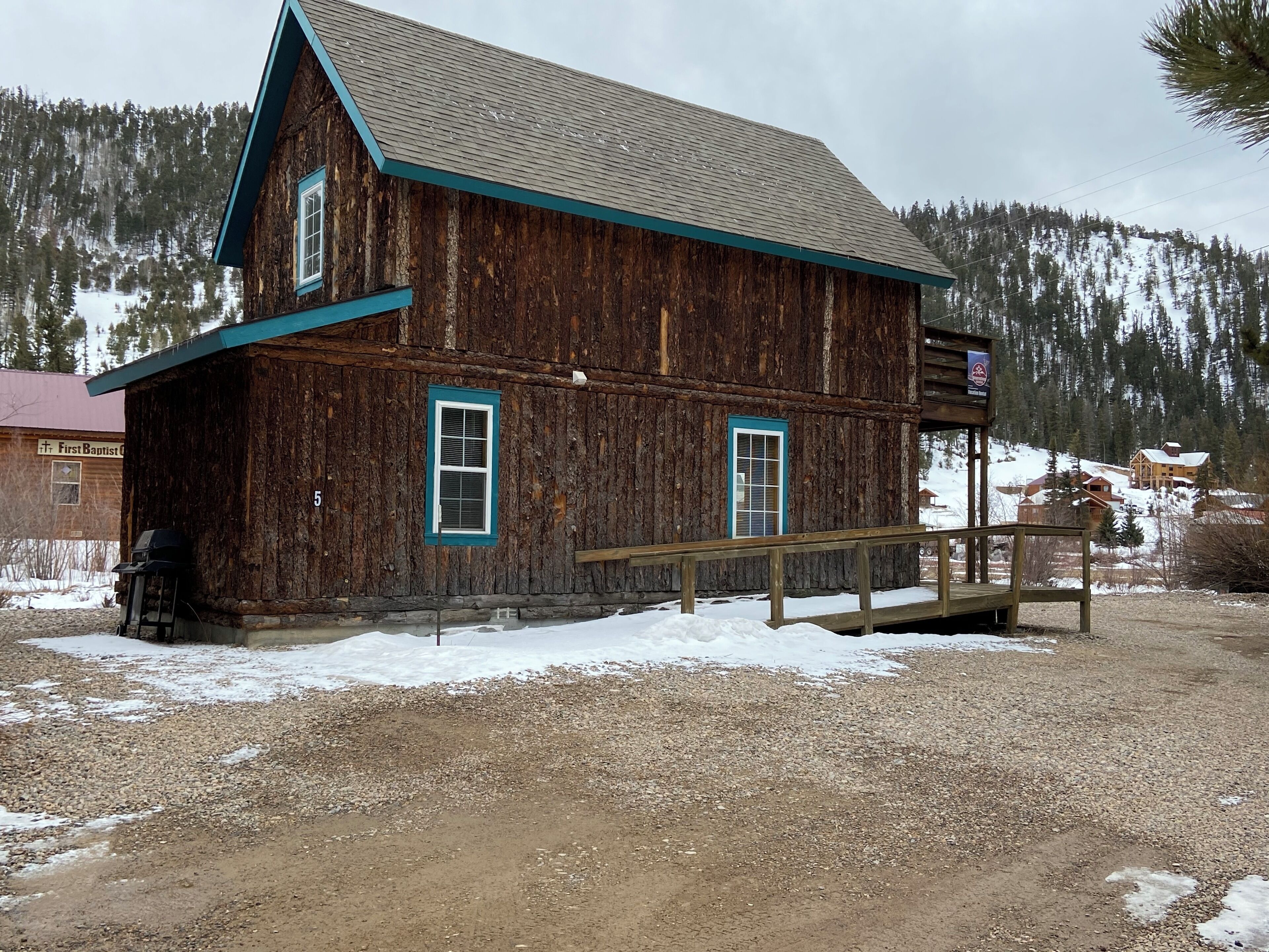 A rustic wooden building with a porch and two windows, surrounded by a gravel area and snow patches.