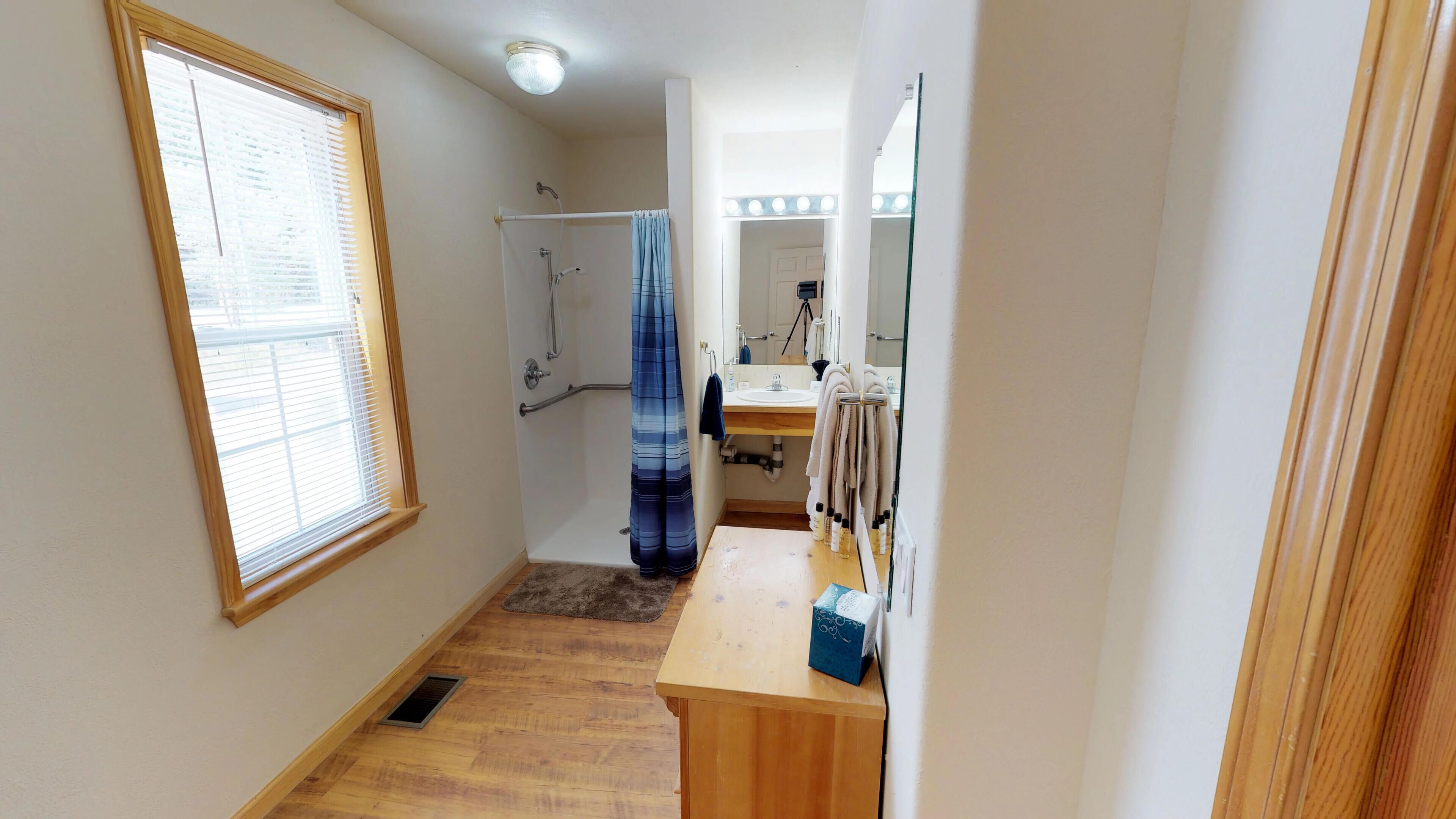 Ein schmaler Korridor mit Holzboden, einem Fenster mit Jalousien, einer Duschkabine mit blauer Duschvorhang und einem Waschtisch mit Spiegel und Beleuchtung.