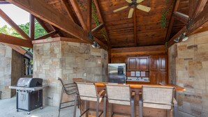 An outdoor kitchen with stone walls, a grill, bar stools, and a ceiling fan.