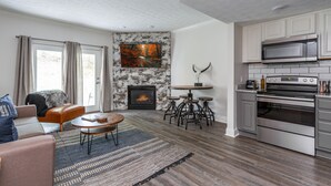 A modern living room with a fireplace, a dining area with stools, and a kitchen with stainless steel appliances.