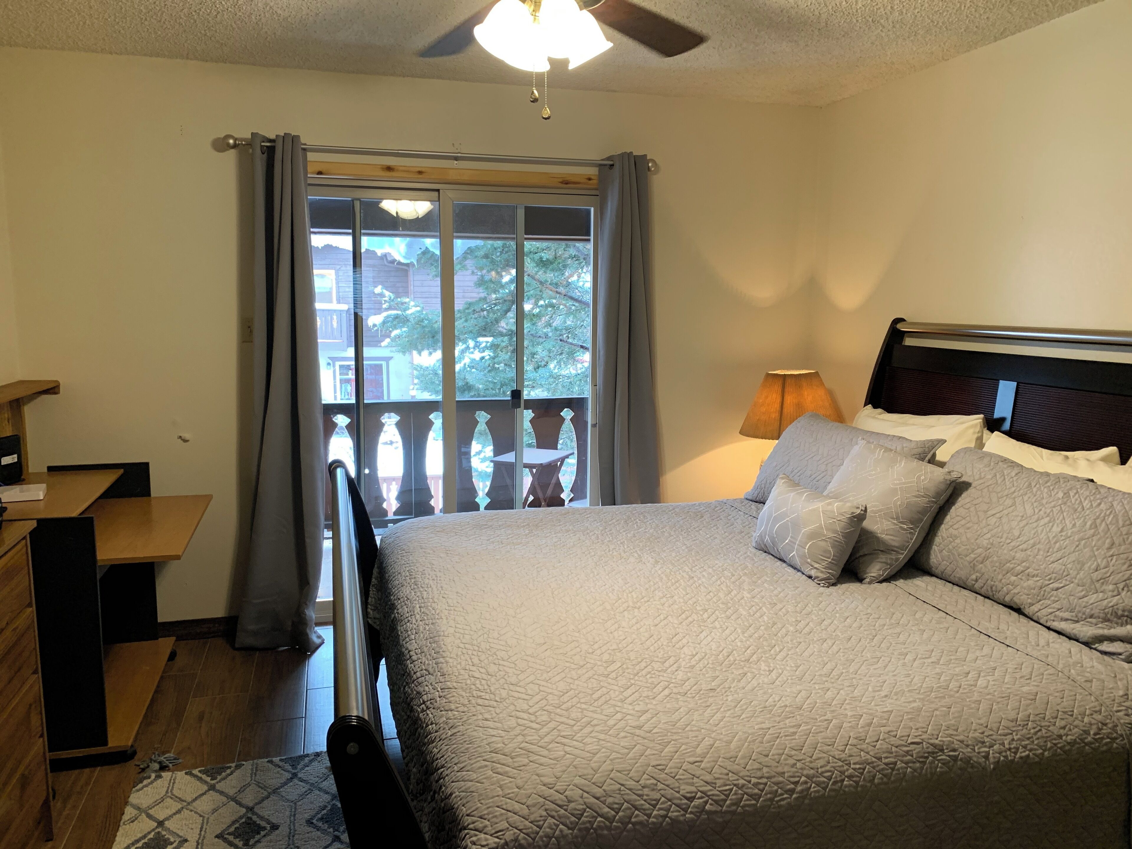 A bedroom with a bed, a desk, a chair, a window with curtains, and a balcony.