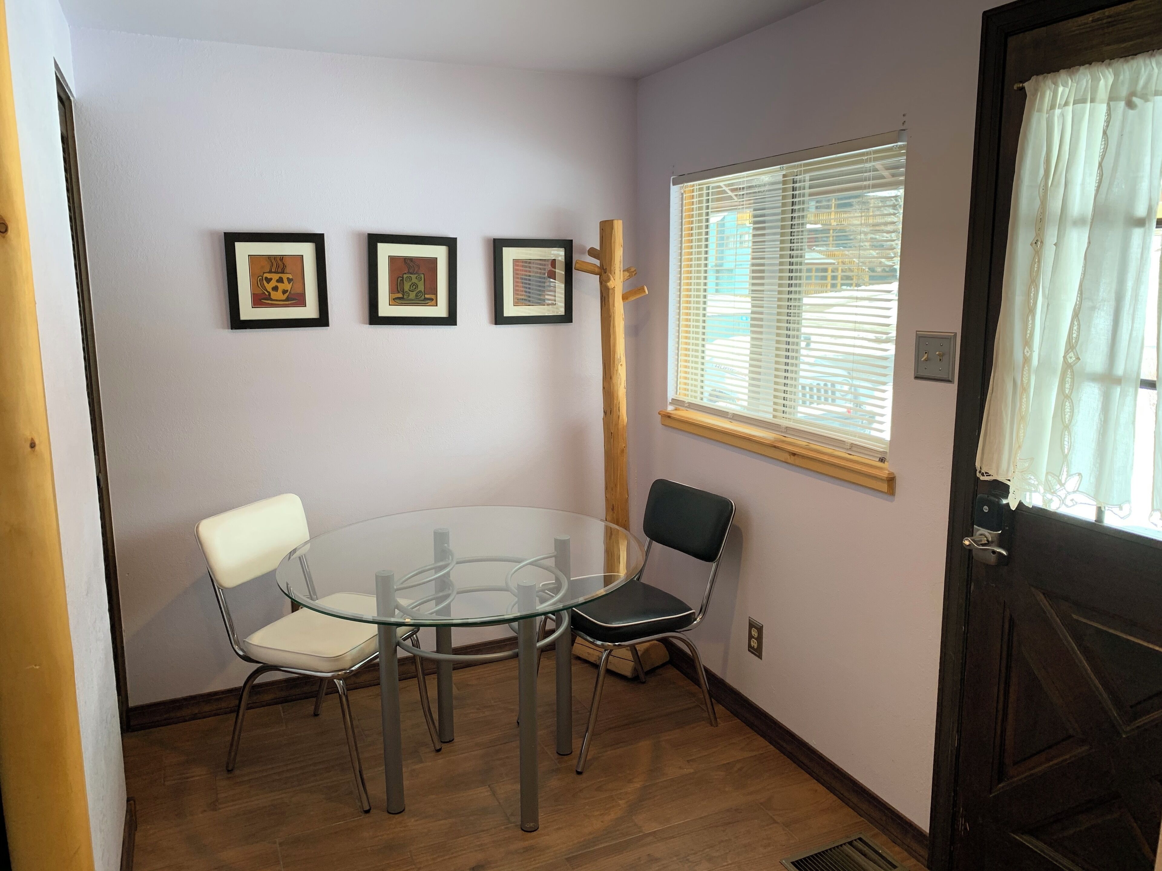 A room with a glass table surrounded by chairs, a window with blinds, and three framed pictures on the wall.