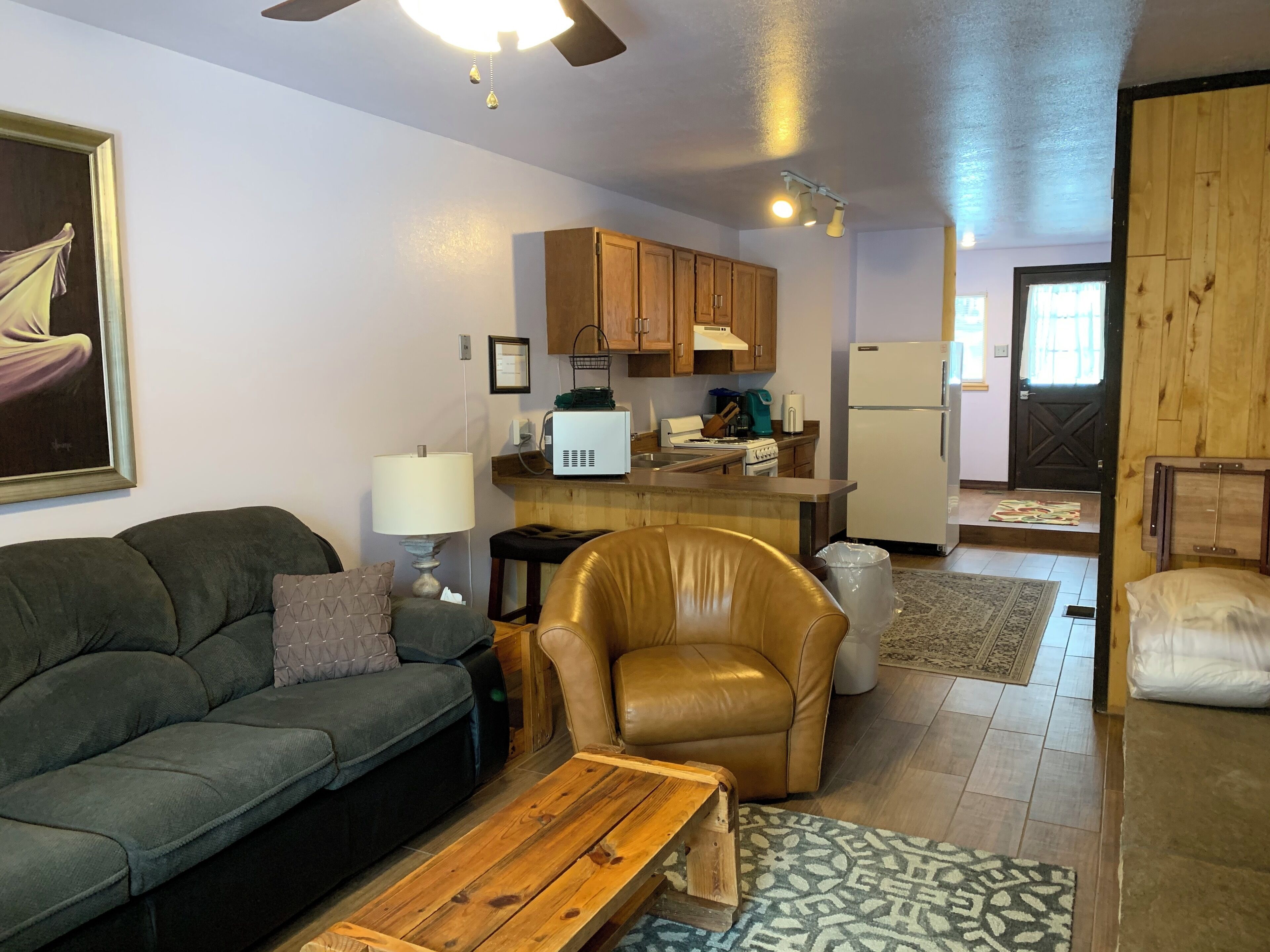 A living room with a sofa, a leather chair, a wooden coffee table, and a kitchen area with wooden cabinets, a stove, and a refrigerator.