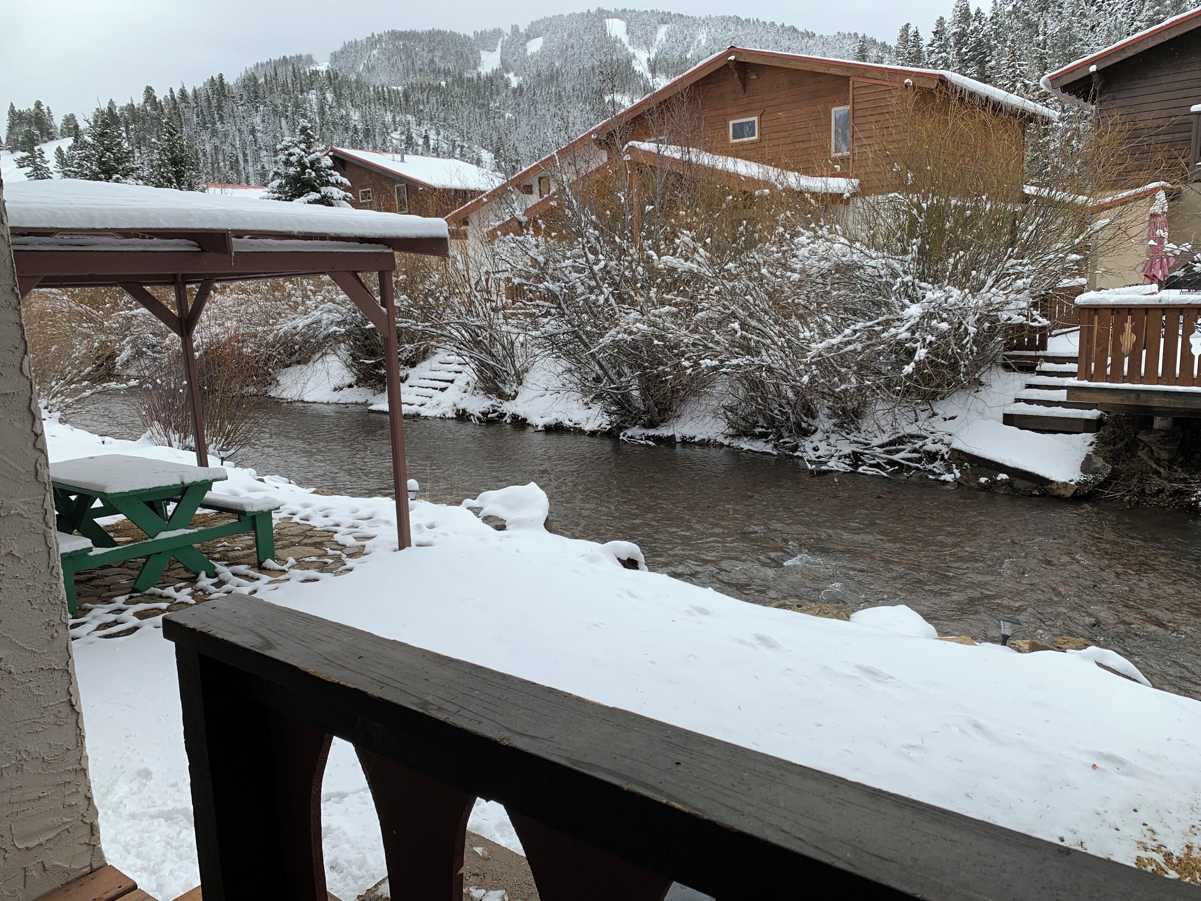 A wooden cabin with a river in the background, surrounded by snow-covered trees and hills.