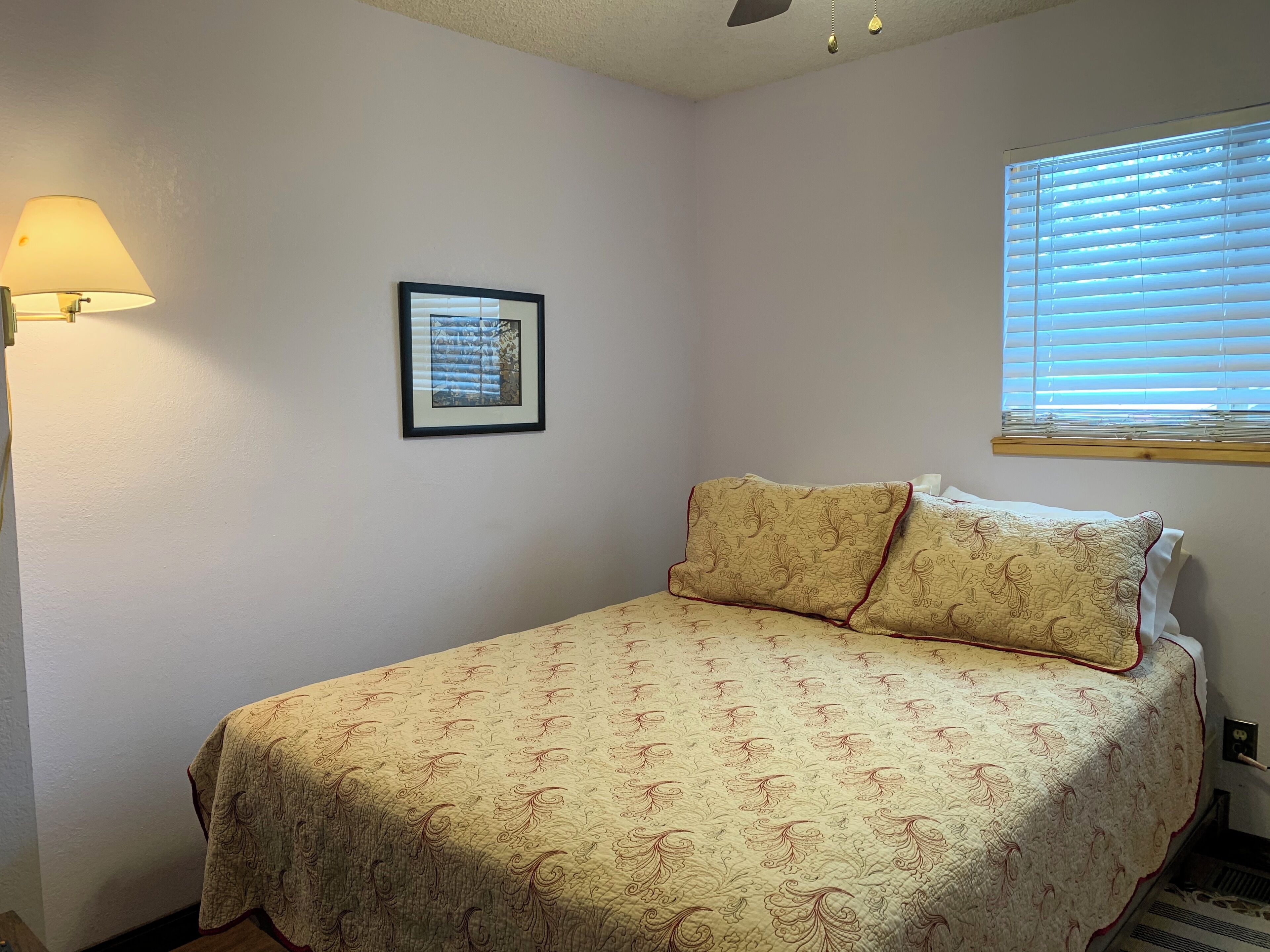 Un dormitorio con cama, una lámpara de pared, un cuadro enmarcado y una ventana con persianas.