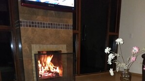 Un salon avec une cheminée, un téléviseur fixé au-dessus, et un vase rempli de fleurs posé sur une table en bois.