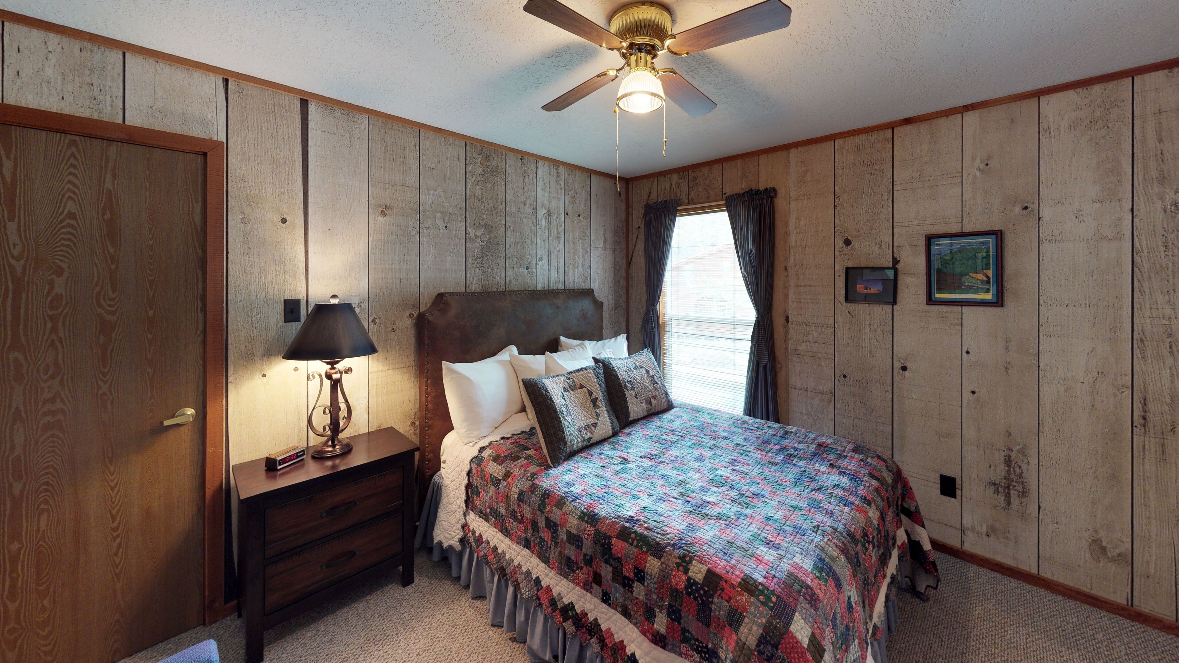 A bedroom with wooden panel walls, a bed with a colorful quilt, a nightstand with a lamp, and a ceiling fan.