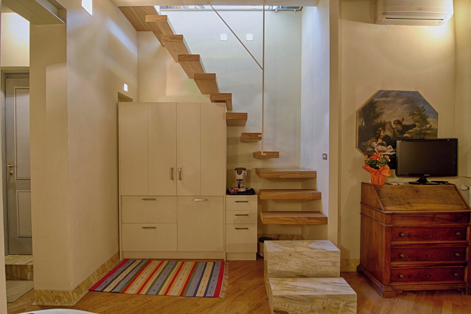Un escalier en bois menant à une mezzanine, une armoire blanche, une machine à café, un téléviseur posé sur un meuble en bois et un tableau accroché au mur.