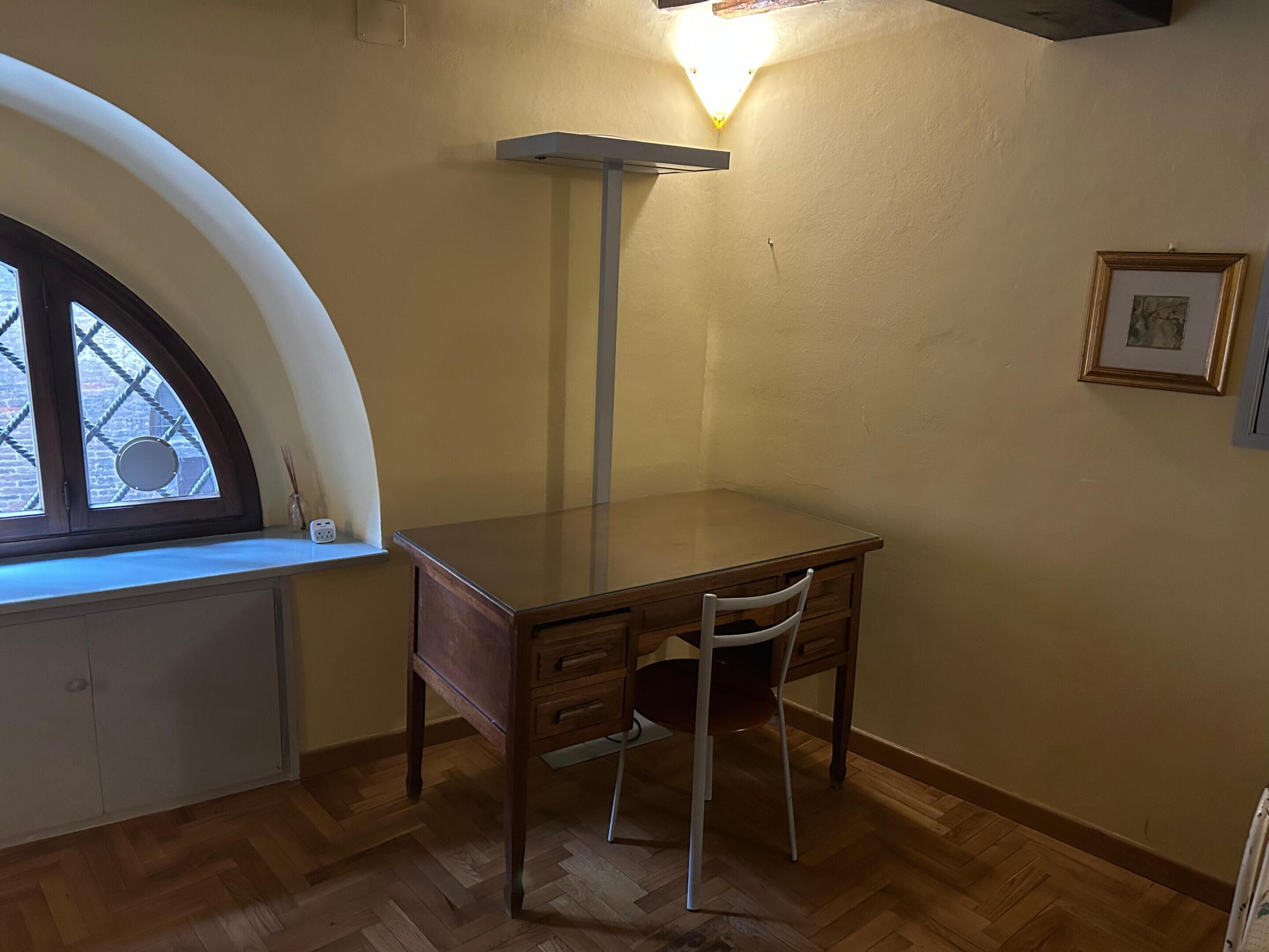 A corner room with a wooden desk, a chair, a radiator, a wall-mounted light fixture, and a framed picture.