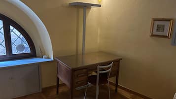 A corner room with a wooden desk, a chair, a radiator, a wall-mounted light fixture, and a framed picture.