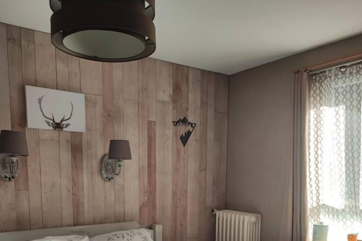 A bedroom with a wooden headboard, a bed with patterned bedding, two wall-mounted lamps, a radiator, and a window with a curtain.
