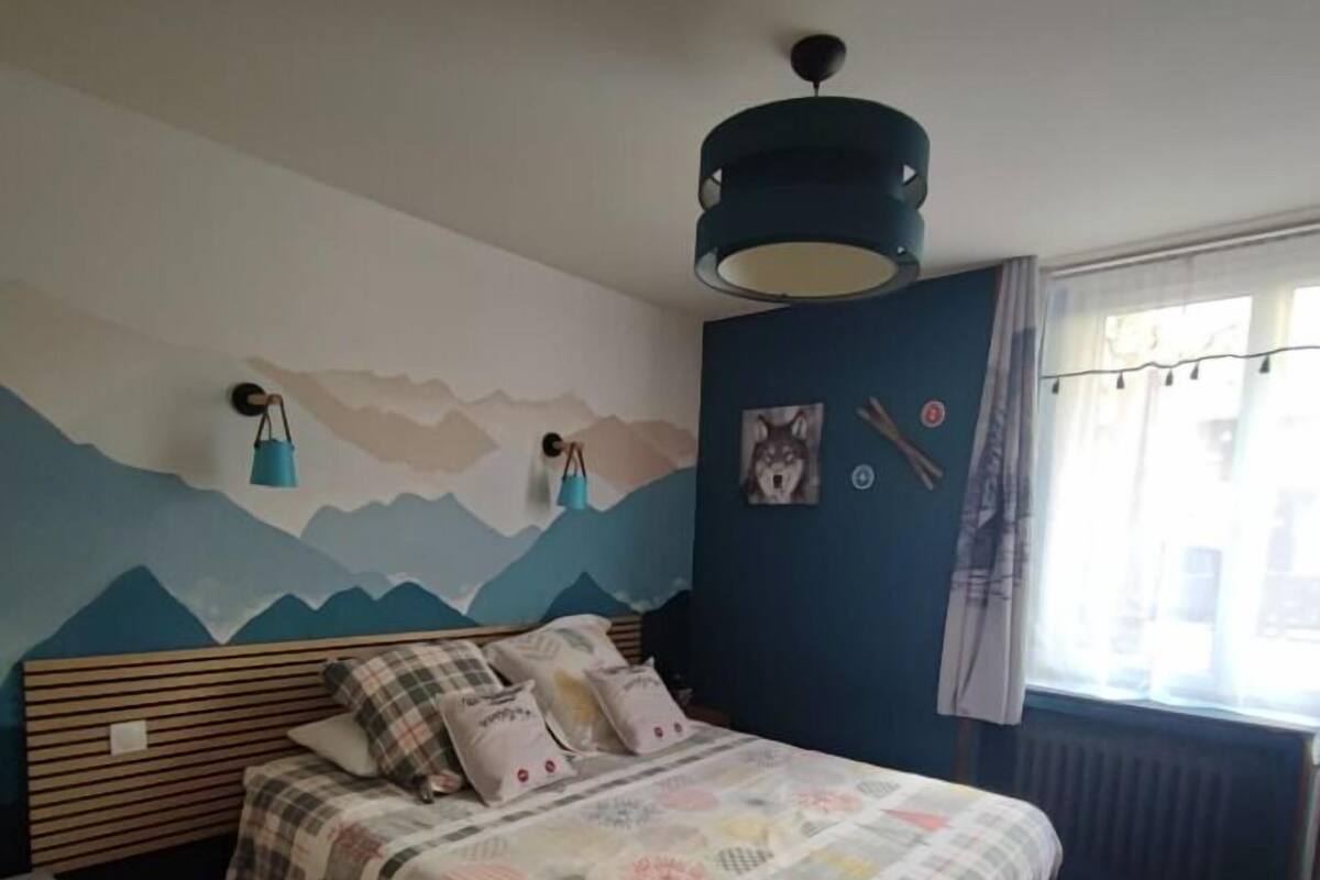 A bedroom with a bed, a wall-mounted lamp, a window with curtains, and a wall with a mountain design.