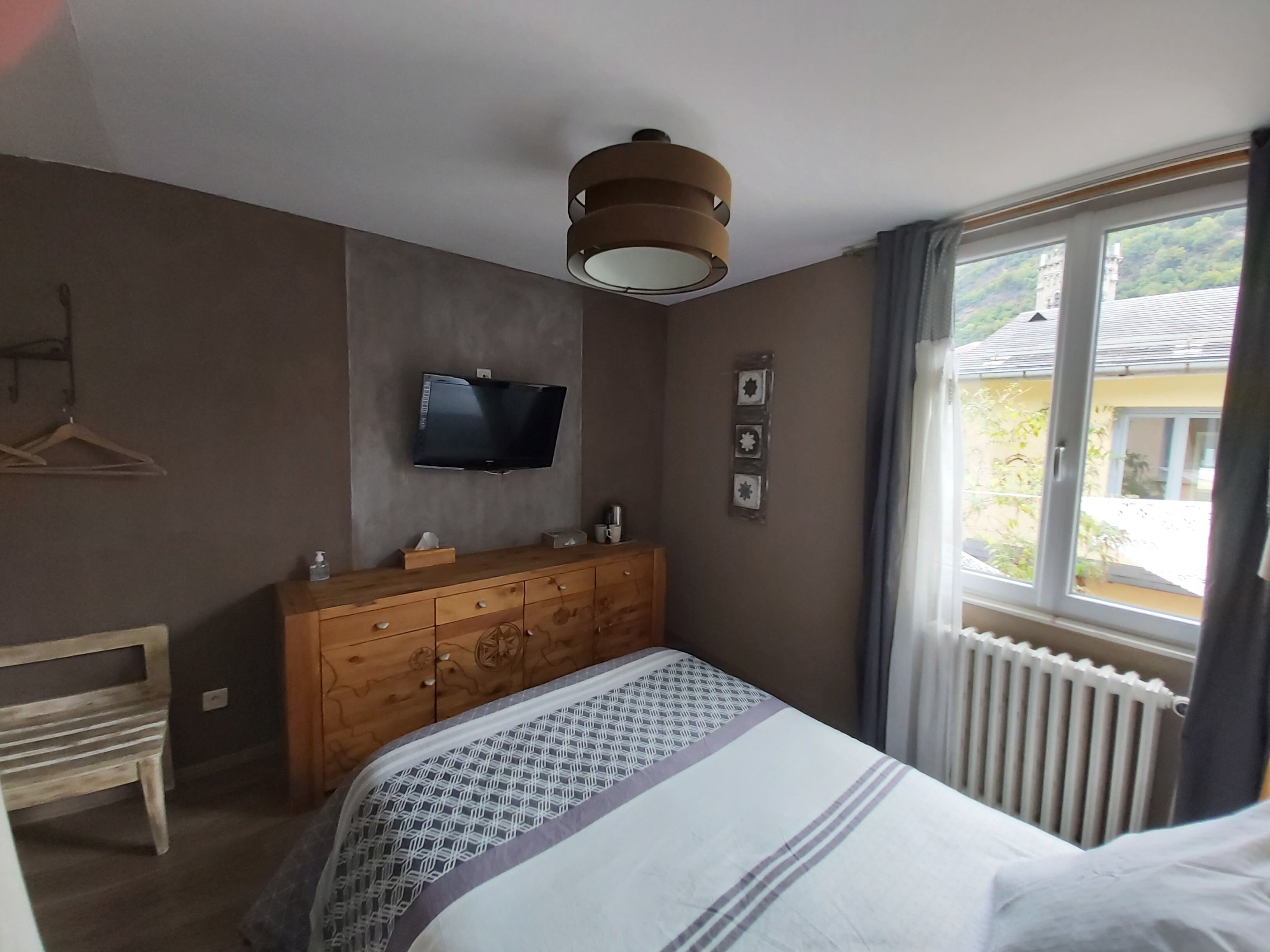 Un dormitorio con cama, cómoda, televisor y ventana con vista a un edificio.