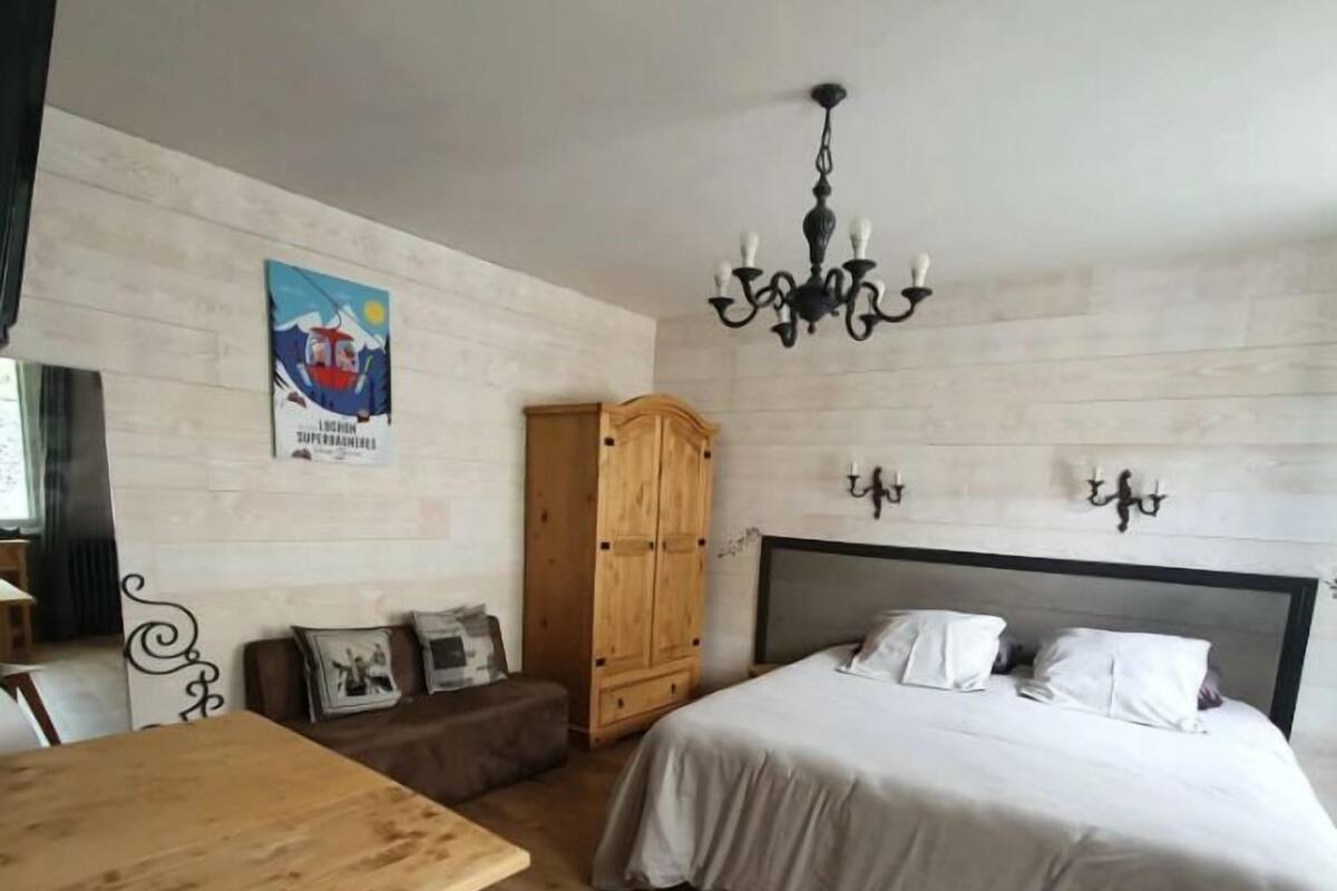 A bedroom with a bed, a wooden wardrobe, a dining table with a chair, and a chandelier.